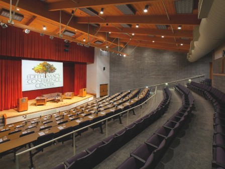 An empty auditorium with rows of seats, a stage with a podium, and a large screen.