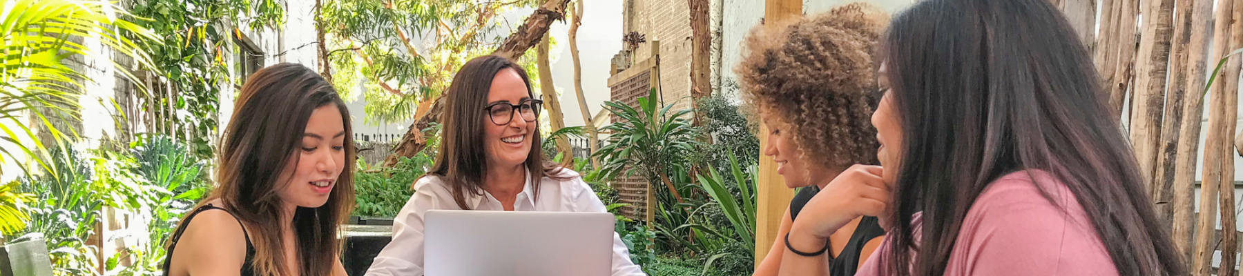 Four people are seated around an outdoor table, engaged in a discussion with notebooks and a laptop. The setting is lush with greenery.